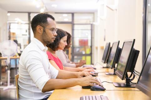 A diverse group of students sitting at individual computer cubicles in a quiet testing environment, focusing on their monitors