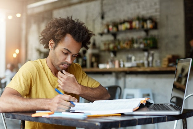 A focused student writing notes in a notebook while referencing a laptop.