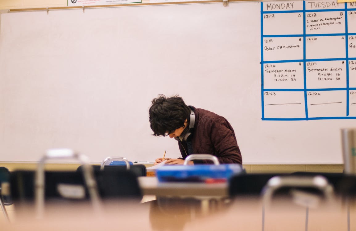 A person sitting alone in a classroom, focused on writing with a whiteboard schedule in the background.