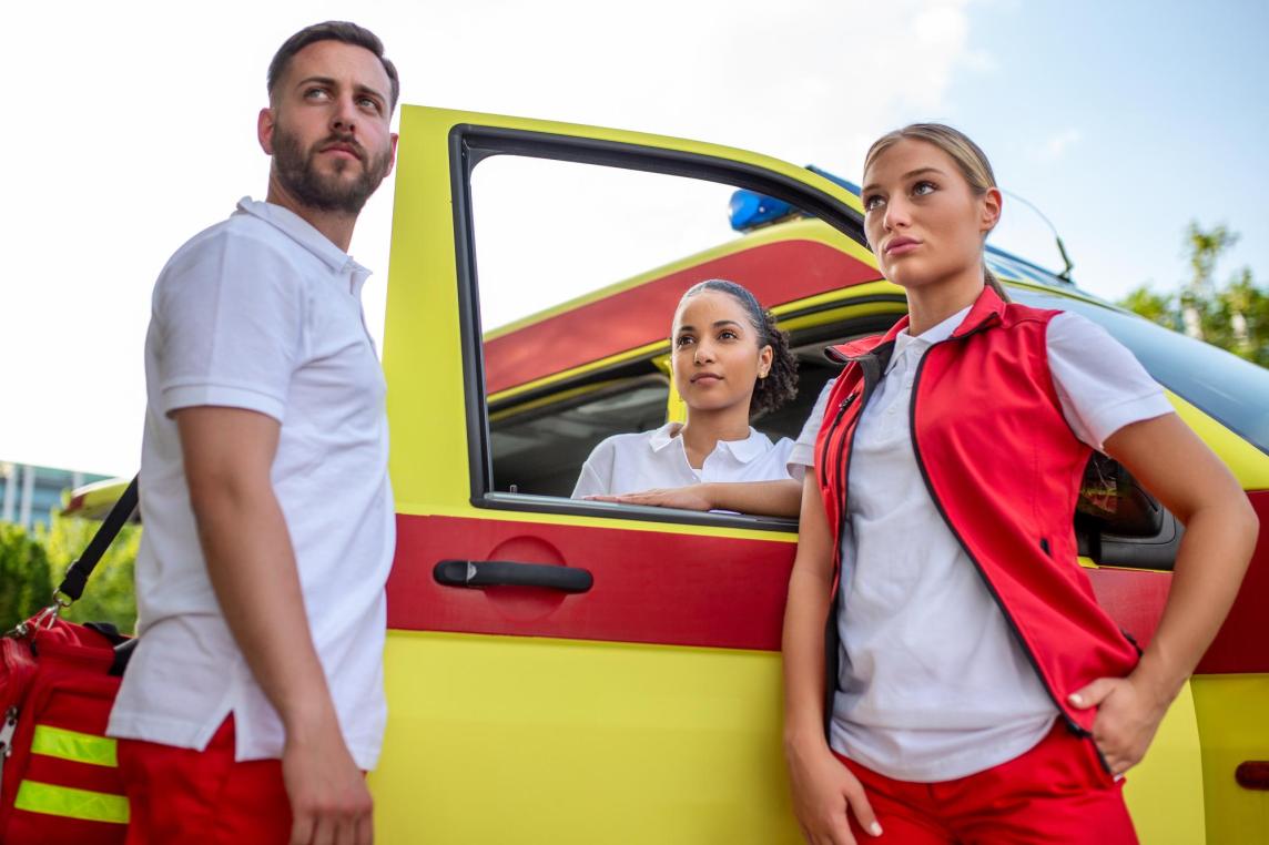 Three paramedics standing in front of an ambulance, one carrying a medical trauma bag, discussing patient care